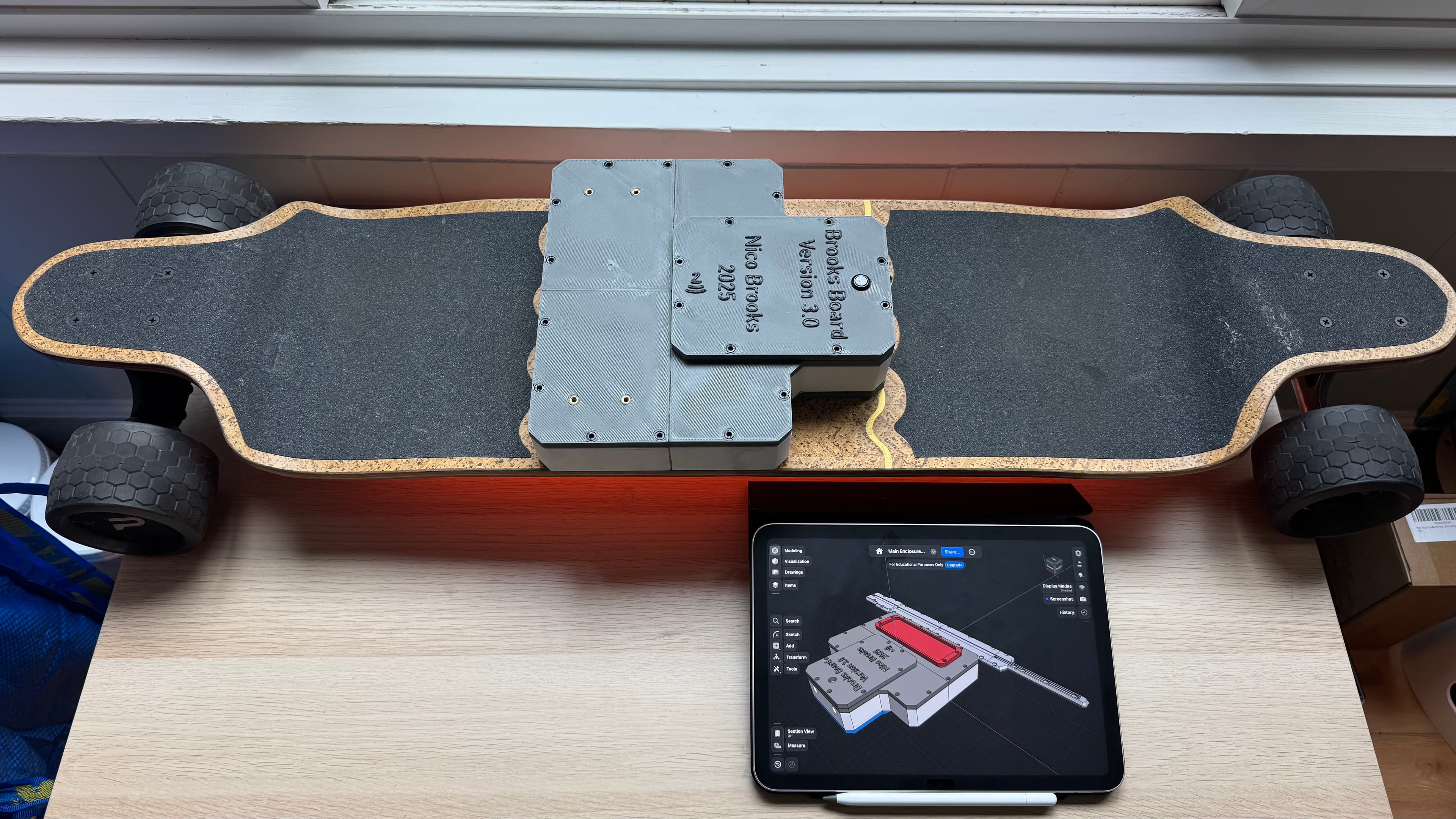BrooksBoard electric longboard with its custom enclosure, beside a tablet displaying the enclosure’s CAD design.